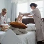 Two housekeepers in uniform fixing a hotel room bed with fresh linen and pillows.